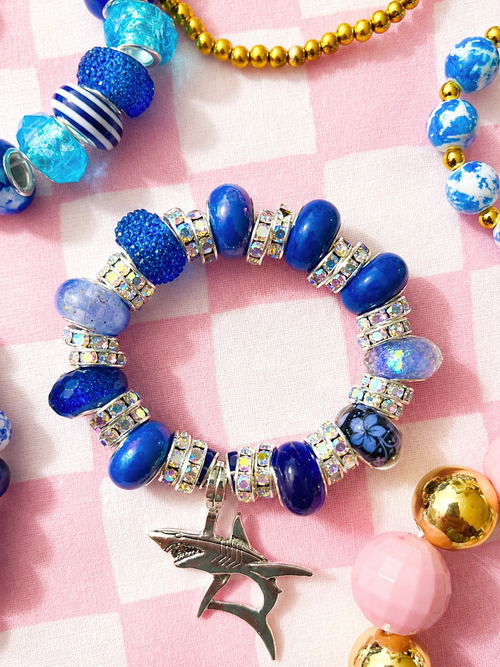 A beaded stretch bracelet featuring royal blue beads, silver rhinestone spacer beads, a blue floral charm, and a silver shark charm, displayed on a pink and white checkered fabric. Ava’s Creations