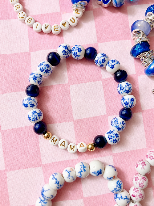 A beaded stretch bracelet arranged in a heart shape on a pink checkered background. The personalized bracelet features white beads with blue floral patterns, solid dark blue beads, small gold spacer beads, and white letter beads spelling 'MAMA'.